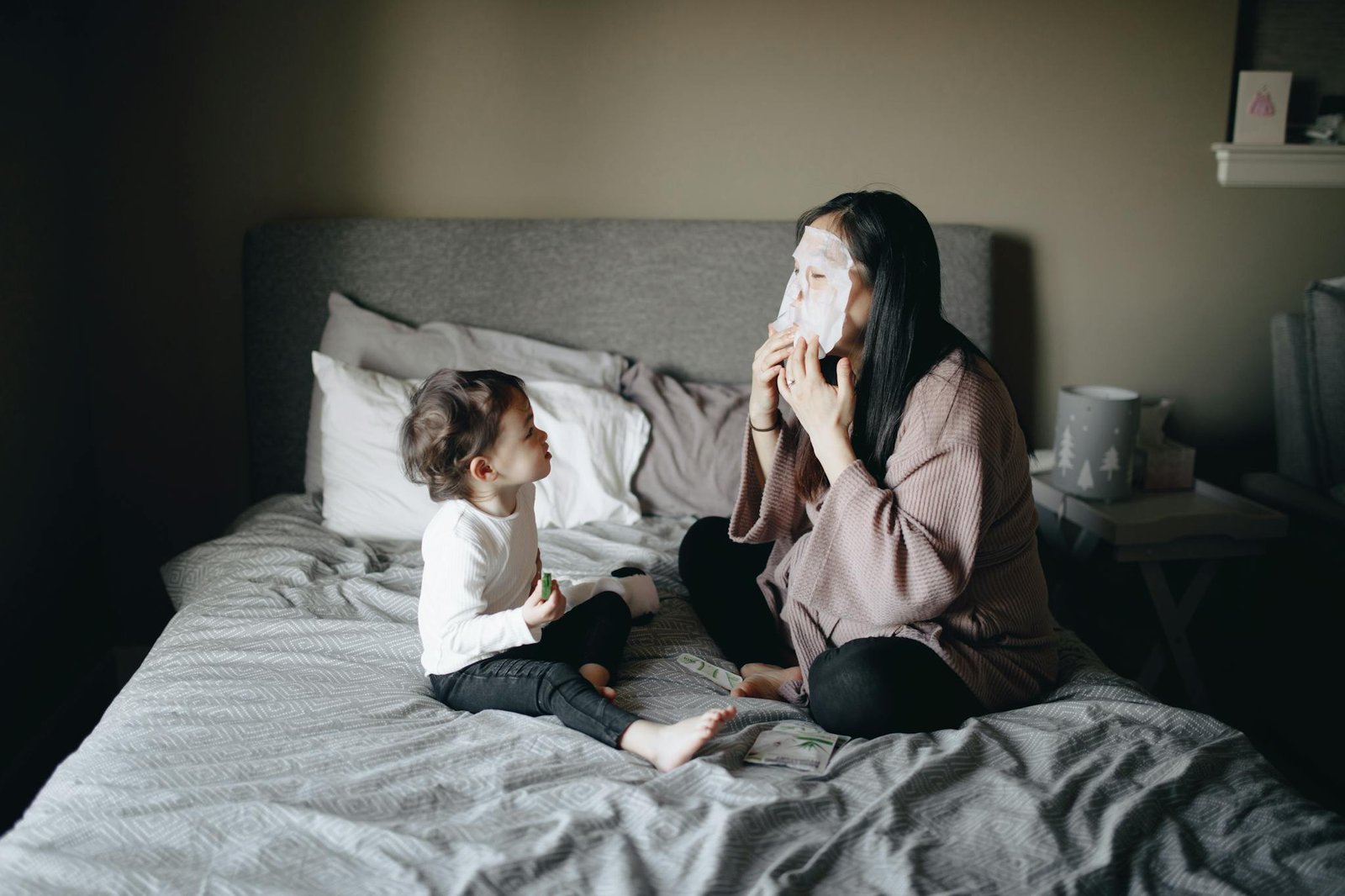 A mother and child bonding over a skincare routine, sitting on a bed indoors.