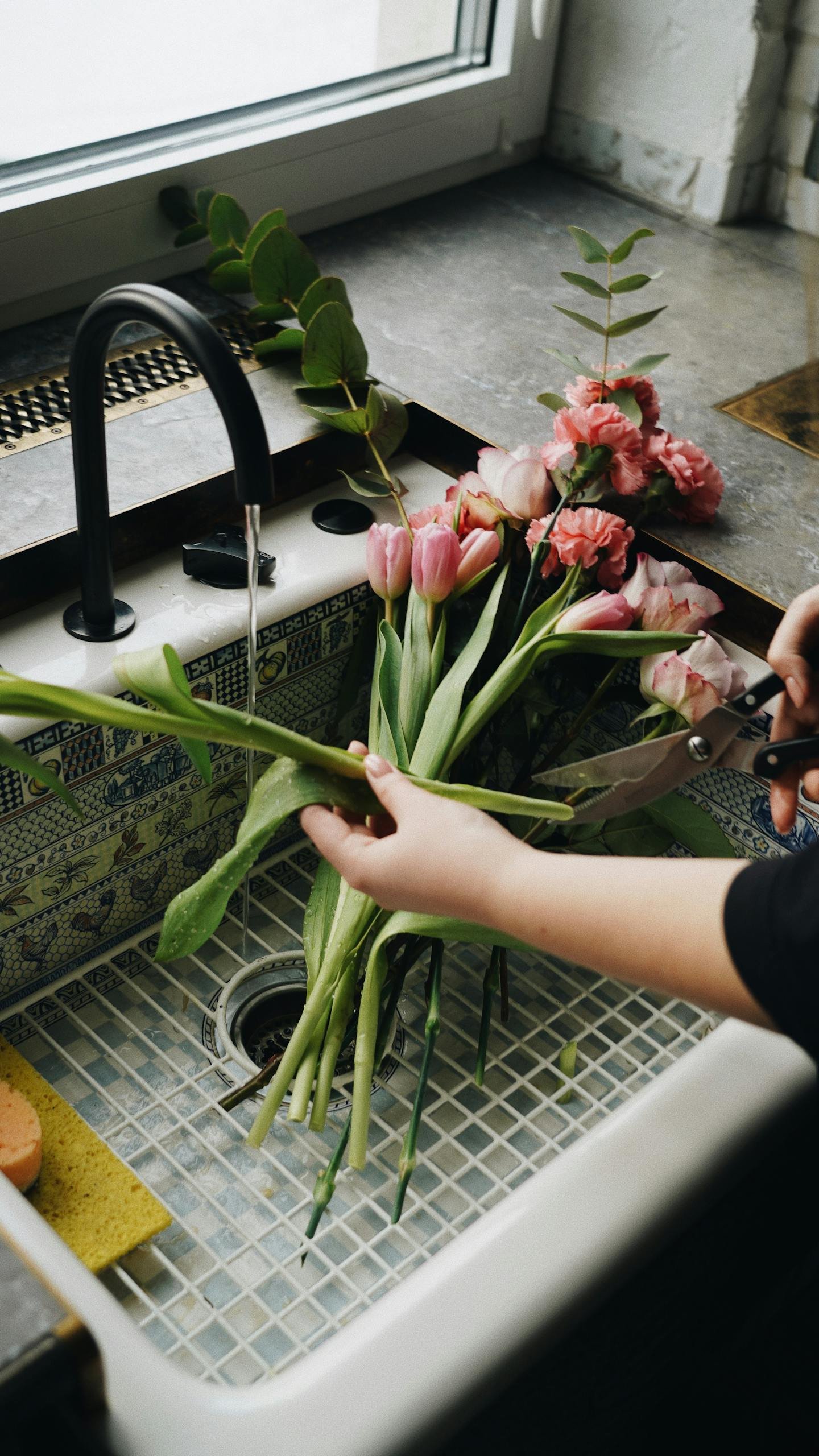 A person trims pink tulips at a kitchen sink, creating a floral arrangement.