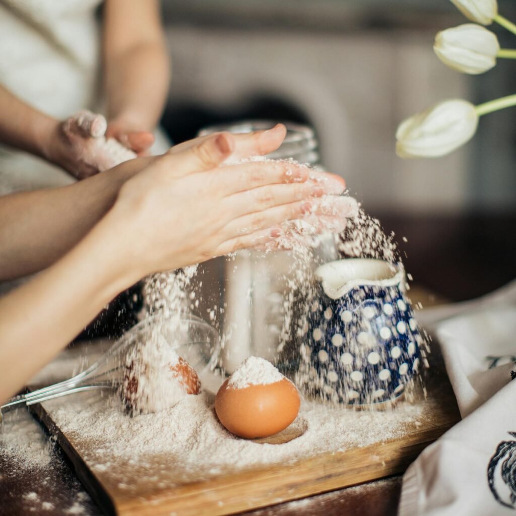 Hands of parent and child baking together, scattering flour on a wooden board with eggs and teapot nearby.